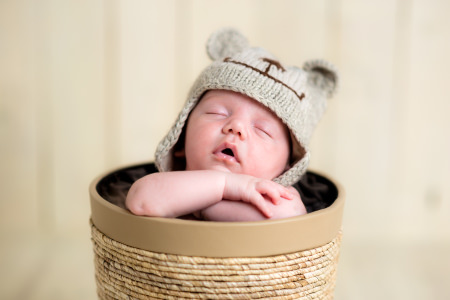 Front view of a cute white Caucasian newborn baby boy sleeping in a decorative basket, crossed hands under the chin holding his head, wearing a hand knitted bear like wool hat. Shot on Canon EOS into a studio, selective focus with sharp peak on baby's face. Warm toned edit to emphasize overall softness feel.